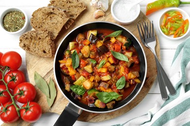 Photo of Frying pan with tasty ratatouille, ingredients and bread on white wooden table, flat lay