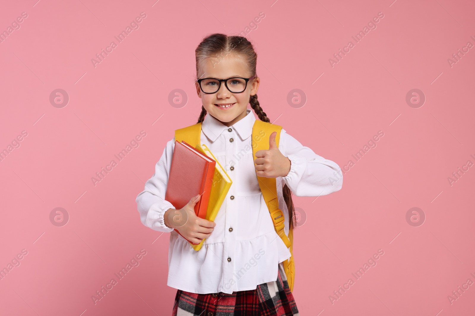 Photo of Happy schoolgirl in glasses with backpack and books showing thumb up gesture on pink background