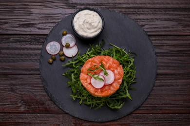 Tasty salmon tartare with radish, capers, arugula and sauce on wooden table, top view