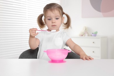 Cute little child eating tasty yogurt from plastic bowl with spoon at white table indoors