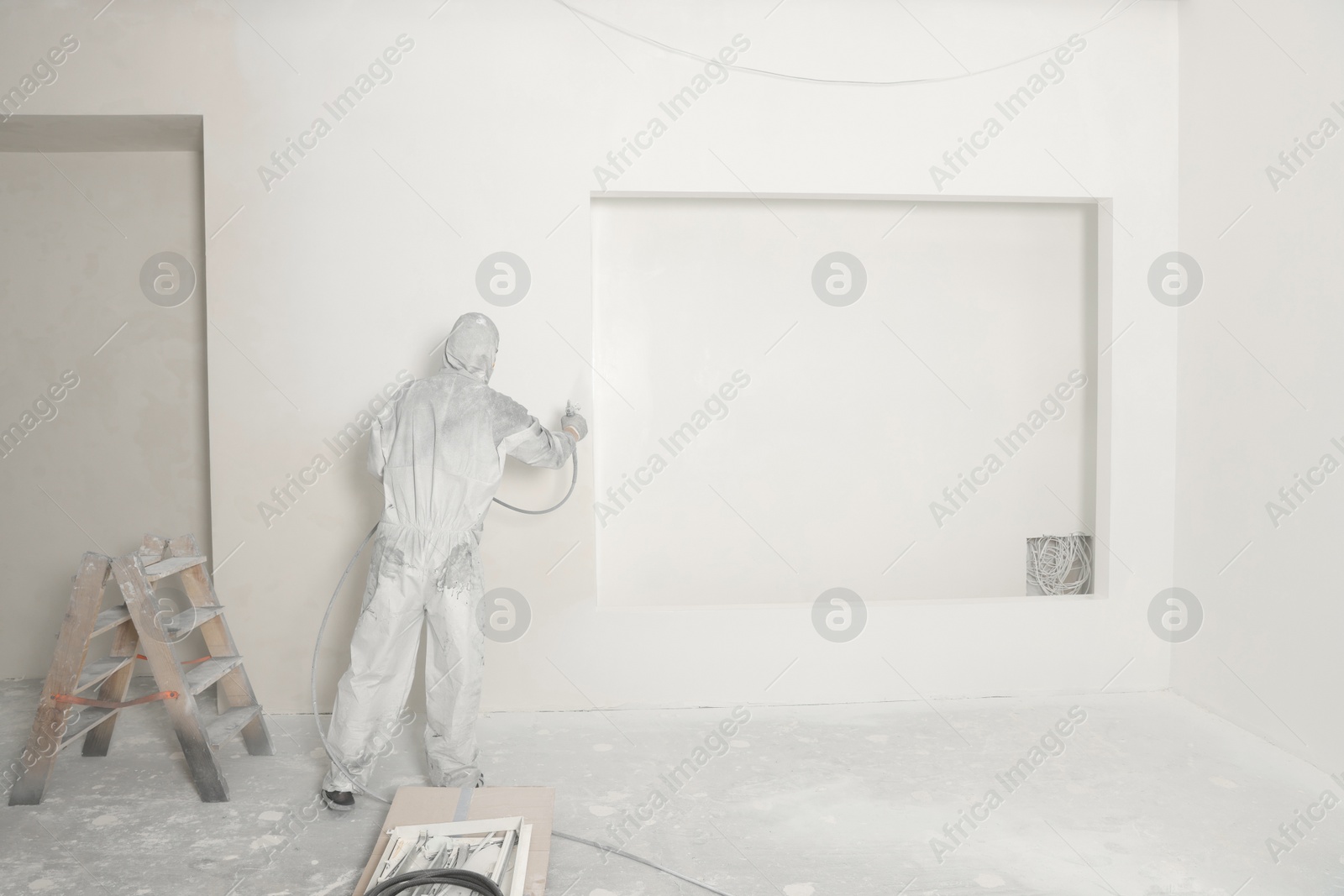 Photo of Decorator in protective overalls painting wall with spray gun indoors