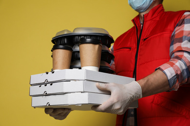 Courier in protective mask and gloves holding order on yellow background, closeup. Food delivery service during coronavirus quarantine