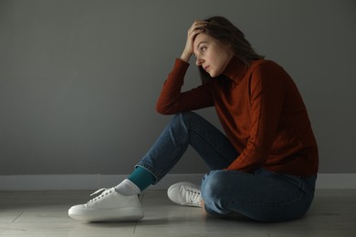 Photo of Sad young woman sitting on floor near grey wall indoors