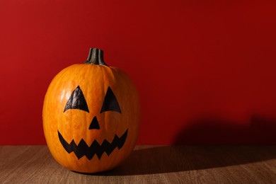 Photo of Pumpkin with drawn spooky face on wooden table against red background, space for text. Halloween celebration