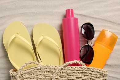 Photo of Wicker beach bag with sunscreen, flip flops and sunglasses on sand, flat lay