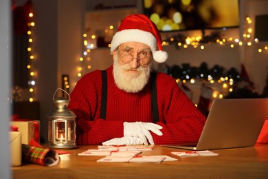 Photo of Santa Claus at his workplace. Letters and laptop on table in room decorated for Christmas