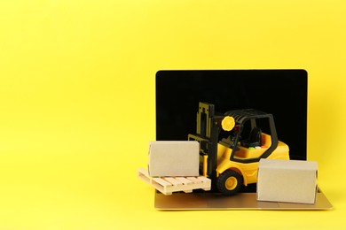 Photo of Laptop, toy forklift with wooden pallet and boxes on yellow background, space for text