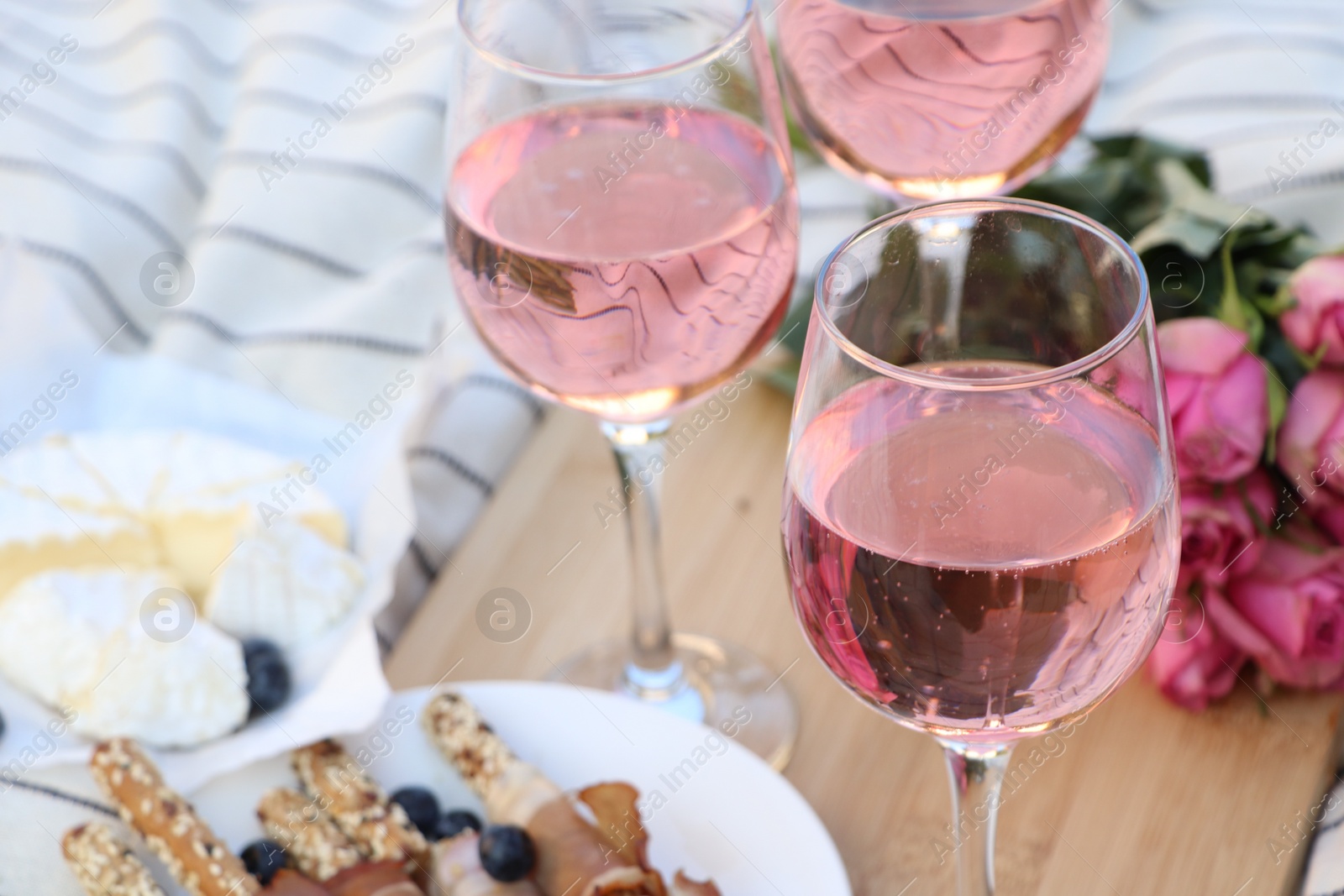 Photo of Glasses of delicious rose wine, flowers and food on white picnic blanket, closeup