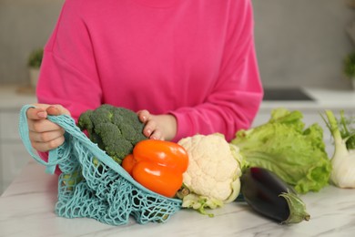 Woman with string bag of fresh vegetables at light marble table, closeup