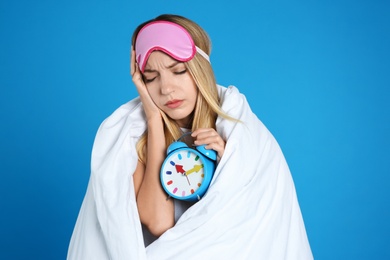 Young woman in sleeping mask wrapped with blanket holding alarm clock on blue background