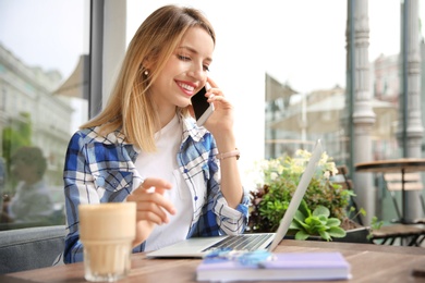 Young woman talking on phone while working with laptop at desk in cafe