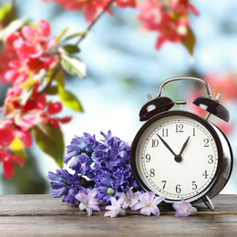 Image of Black alarm clock and flowers on wooden table against blurred background. Spring time