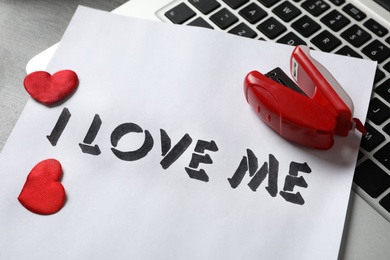 Photo of Paper with phrase I Love Me, laptop, red hearts and stapler on desk, closeup