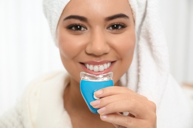 Young African-American woman using teeth whitening device on light background