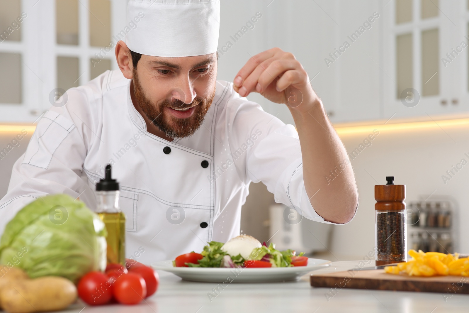 Photo of Professional chef salting delicious salad at marble table in kitchen