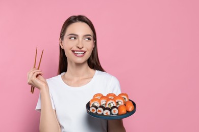 Happy young woman with plate of sushi rolls and chopsticks on pink background