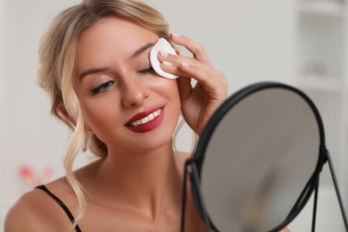 Smiling woman removing makeup with cotton pad in front of mirror indoors, closeup
