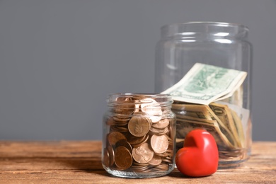 Photo of Red heart and donation jars with money on wooden table against grey background. Space for text