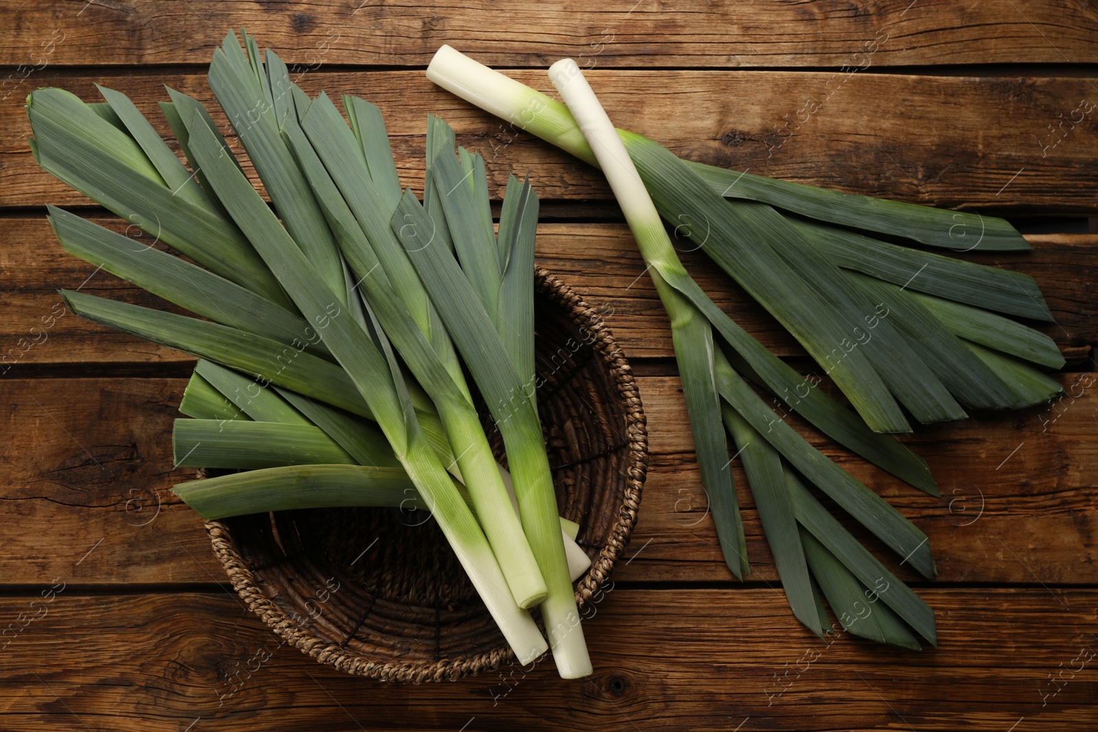Photo of Fresh raw leeks and wicker bowl on wooden table, flat lay