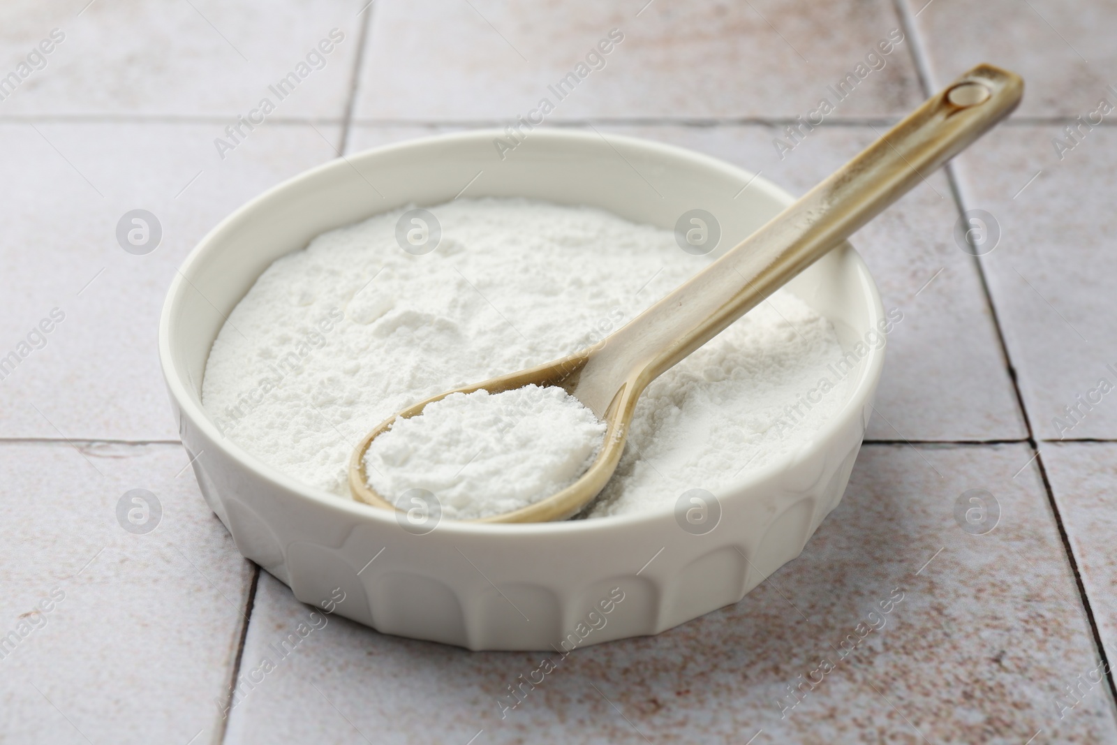 Photo of Baking powder in bowl and spoon on light tiled table, closeup