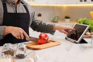 Cooking process. Man using tablet while cutting fresh bell pepper at white marble countertop in kitchen, closeup