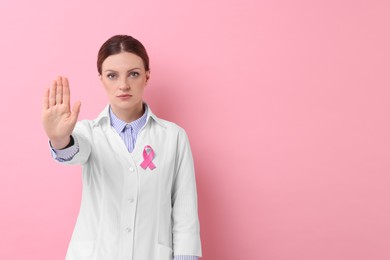 Mammologist with pink ribbon showing stop gesture against color background, space for text. Breast cancer awareness
