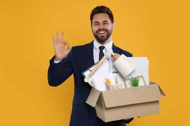 Photo of Happy unemployed man with box of personal office belongings showing ok gesture on orange background