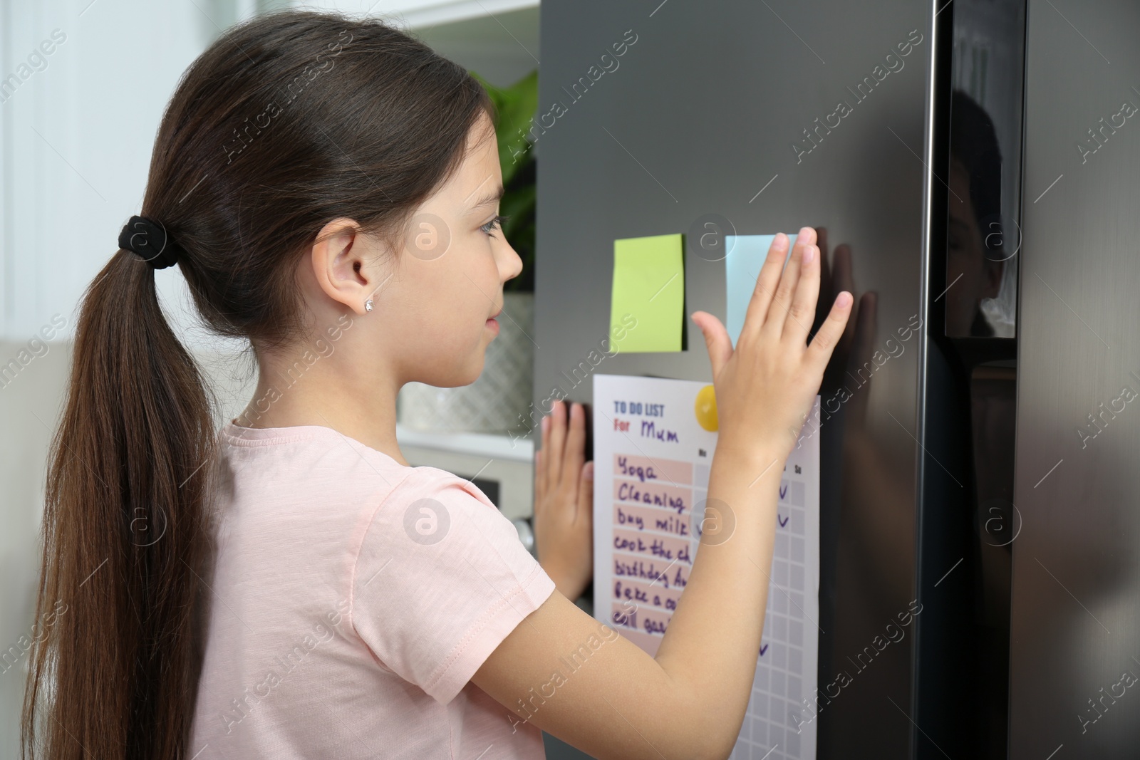 Photo of Little girl putting sticky note near to do list on fridge in kitchen