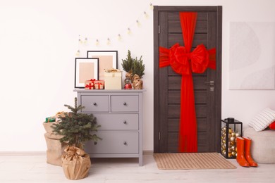 Photo of Wooden door with beautiful red bow near chest of drawers, evergreen trees and gift boxes indoors. Christmas decoration