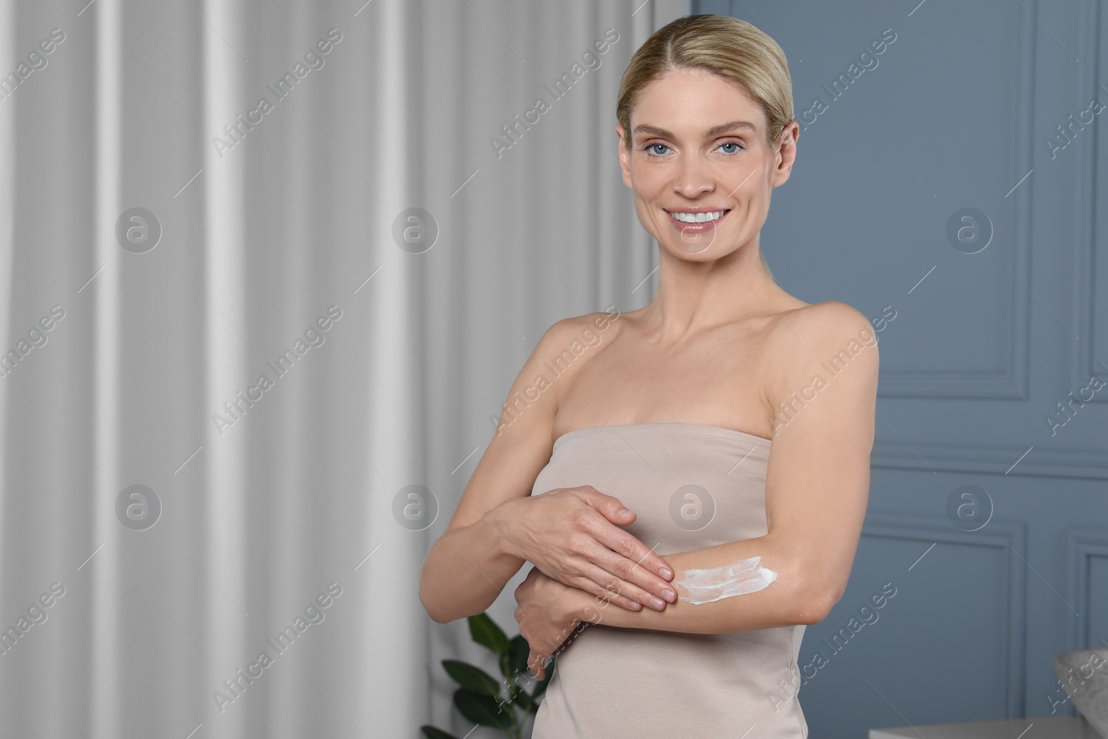 Photo of Happy woman applying body cream onto arm indoors, space for text