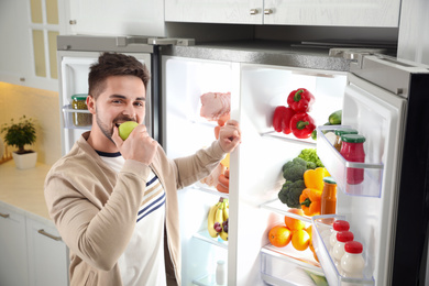 Photo of Young man eating apple near open refrigerator in kitchen