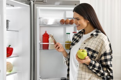 Photo of Young woman with apple and smartphone near modern refrigerator indoors