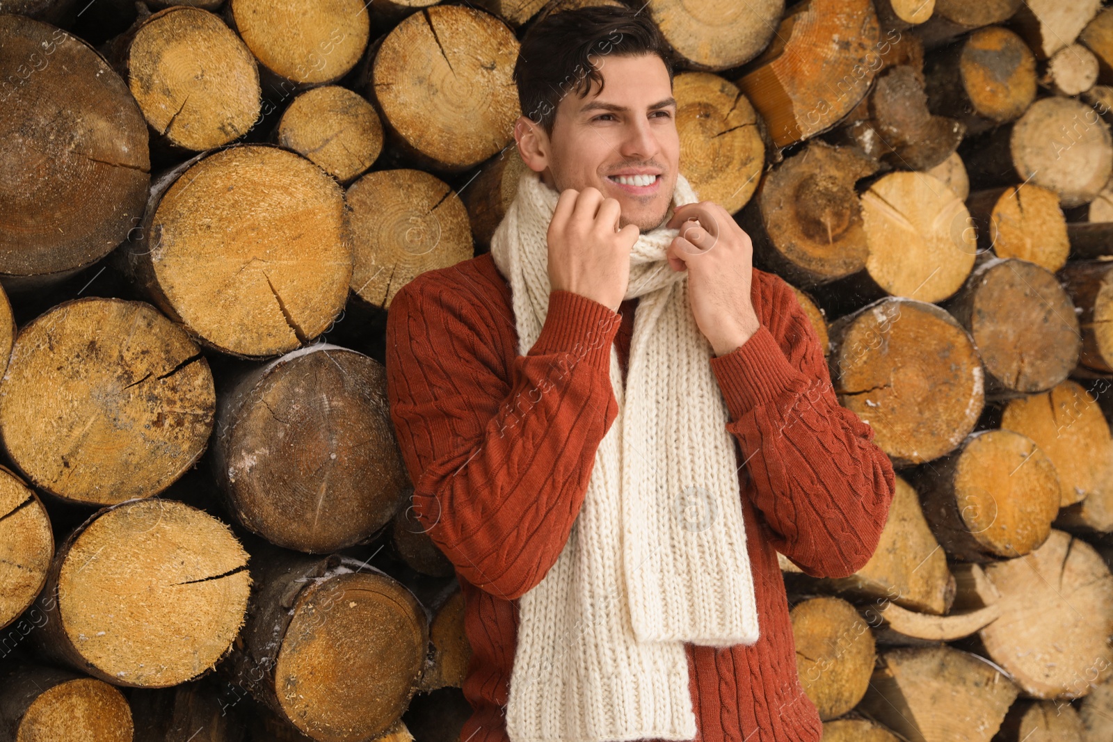 Photo of Handsome man wearing warm sweater and scarf near stack of firewood. Winter season