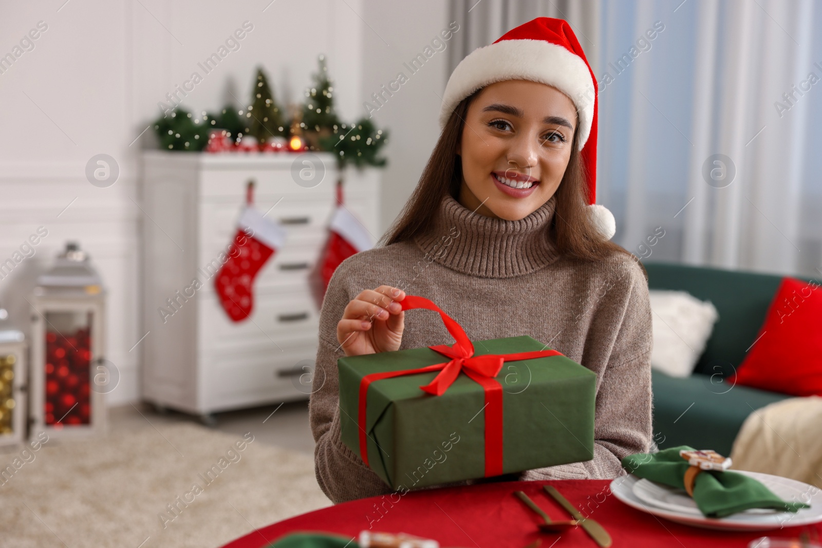 Photo of Happy young woman in Santa hat opening Christmas gift at served table in decorated room