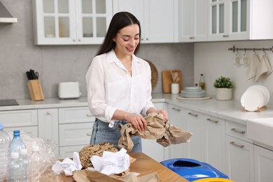 Photo of Garbage sorting. Smiling woman throwing crumpled paper into trash bin in kitchen