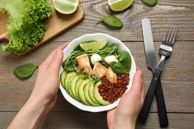 Photo of Woman holding bowl of delicious avocado salad with chicken at wooden table, top view