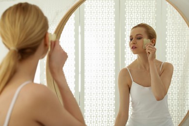 Beautiful young woman doing facial massage with gua sha tool in front of mirror at home