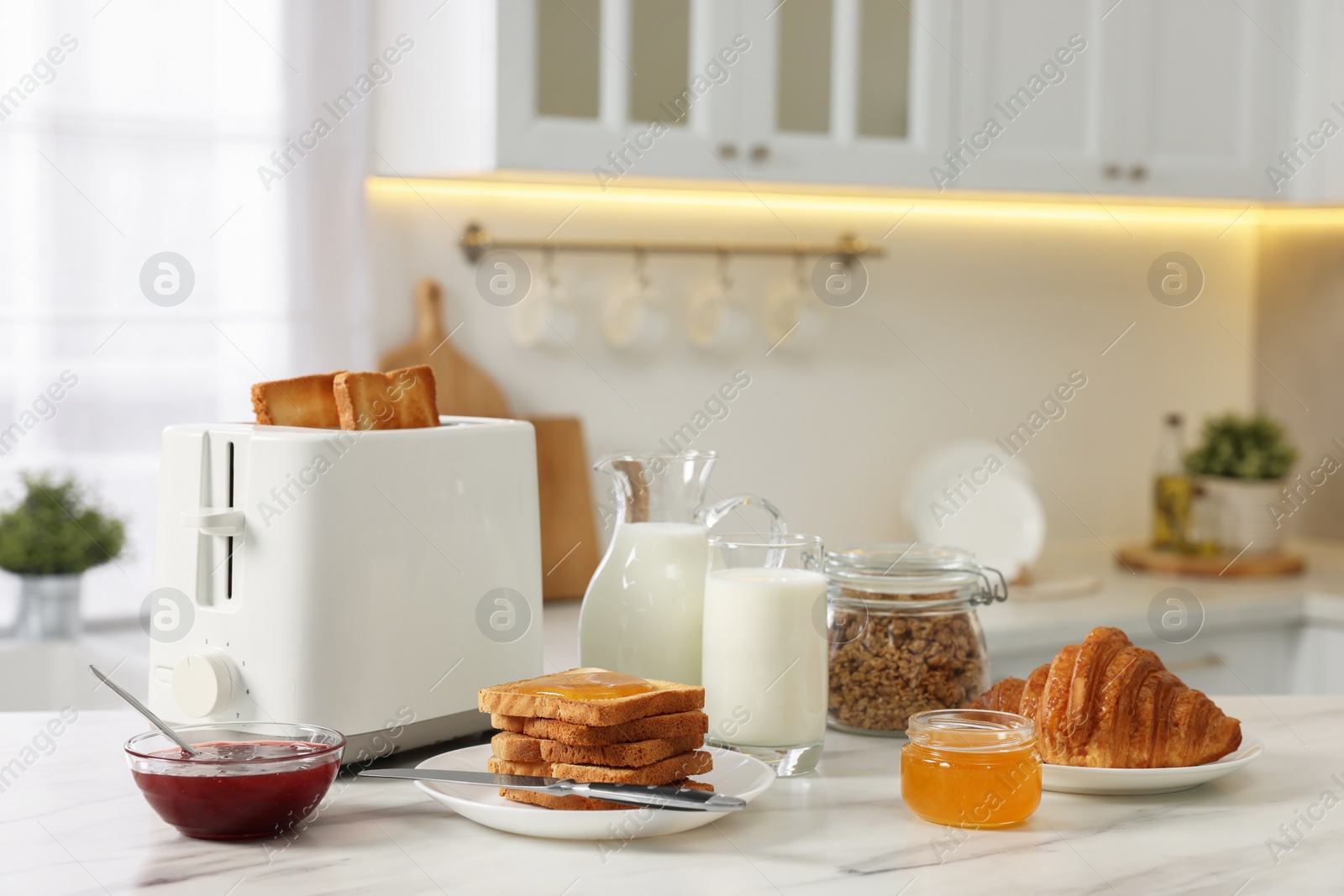 Photo of Making toasts for breakfast. Appliance, crunchy bread, honey, jam, milk and croissant on white marble table in kitchen