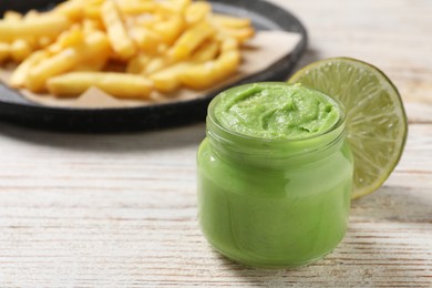 Photo of Jar of avocado dip, lime and plate with french fries on white wooden table, space for text