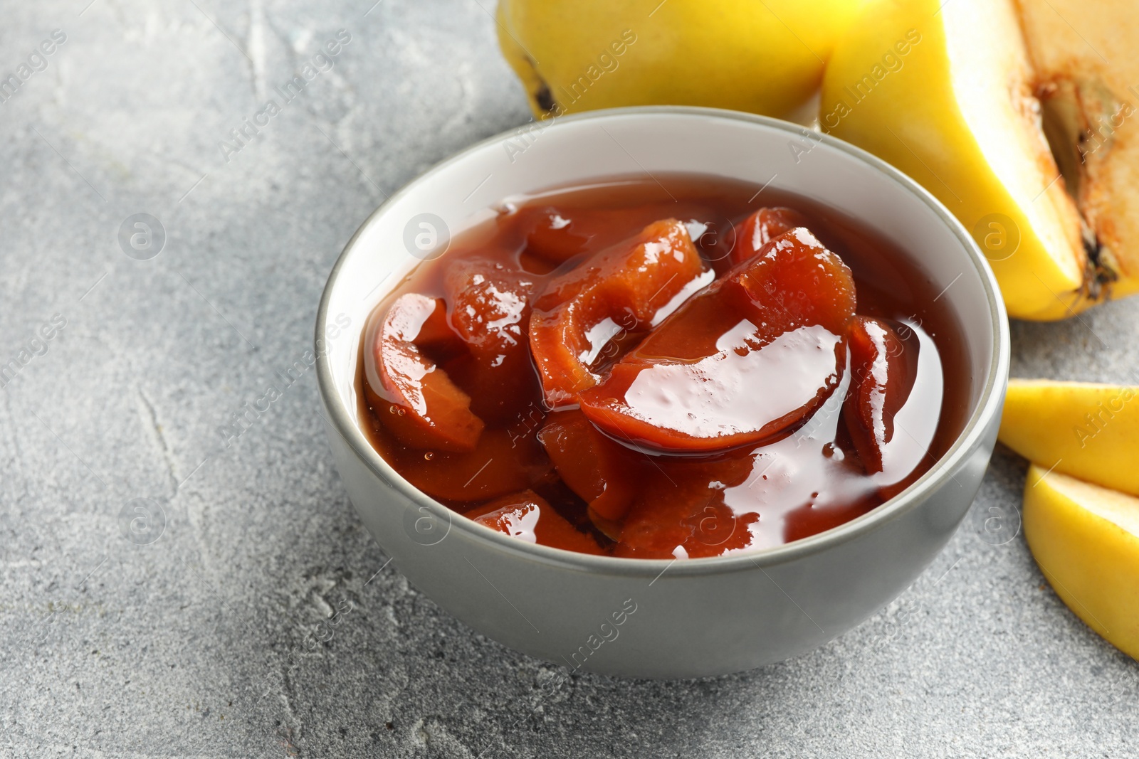 Photo of Tasty homemade quince jam in bowl and fruits on grey textured table, closeup. Space for text