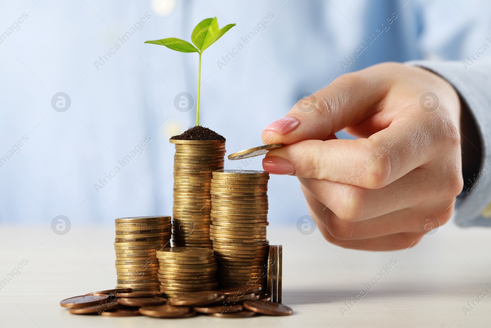 Photo of Woman putting coin onto stack with green sprout at white table, closeup. Investment concept