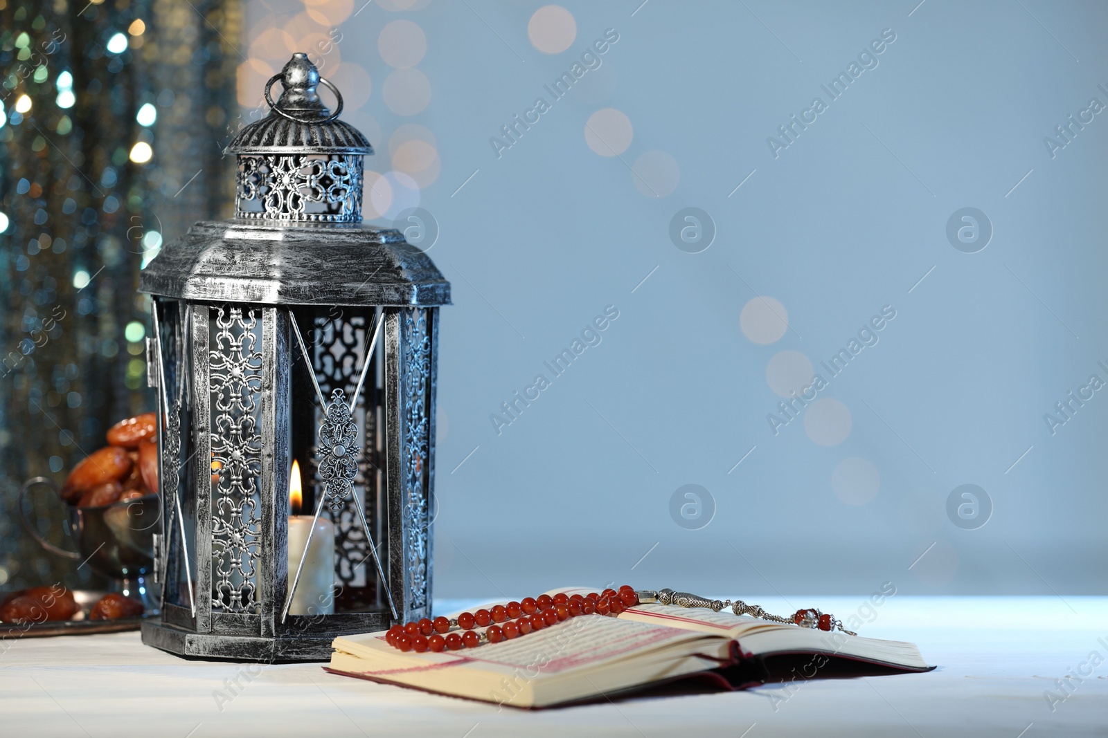 Photo of Arabic lantern, Quran, misbaha and dates on table against blurred lights, space for text