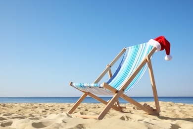 Photo of Deck chair with Santa Claus hat on sandy beach. Christmas vacation