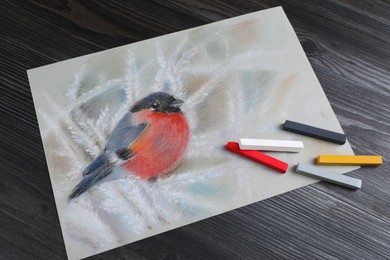 Photo of Colorful chalk pastels and beautiful painting of bullfinch on black wooden table, above view