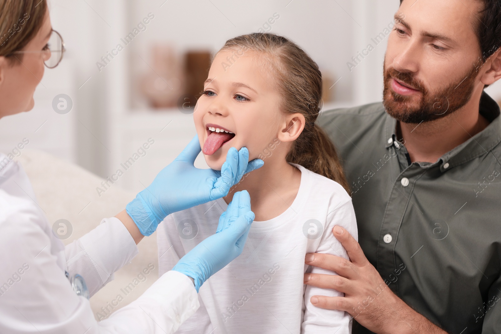 Photo of Doctor examining girl`s oral cavity near her father indoors