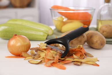 Peels of fresh vegetables and peeler on white wooden table, closeup