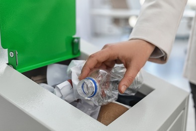 Woman putting used plastic bottle into trash bin in modern office, closeup. Waste recycling