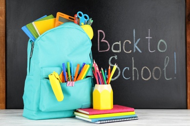 Bright backpack with school stationery on white wooden table near black chalkboard. Back to School