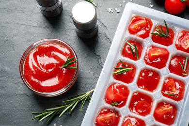 Ice cube tray with tomatoes, sauce and fresh rosemary on grey table, flat lay