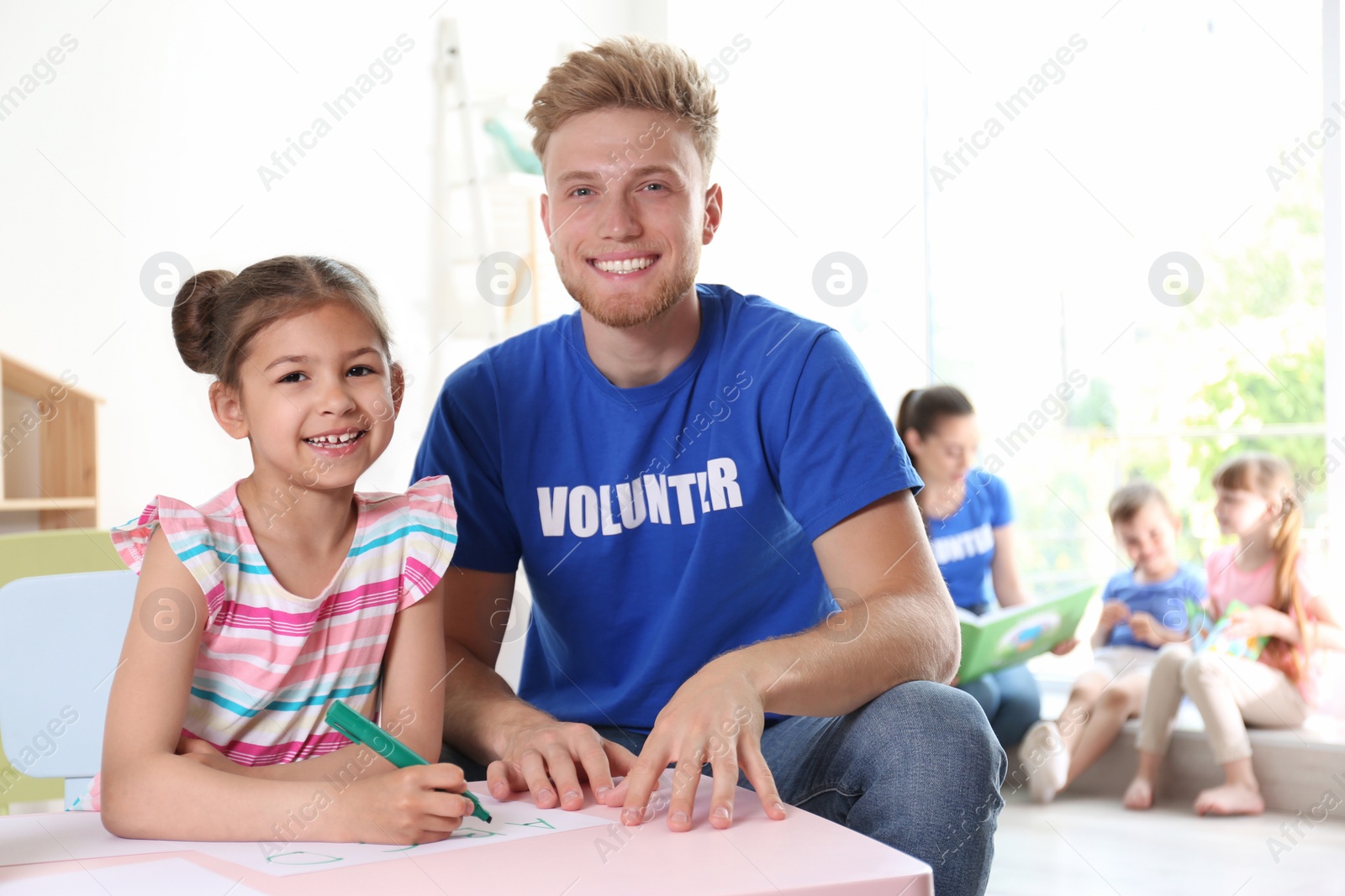 Photo of Little girl learning alphabet with volunteer at table indoors